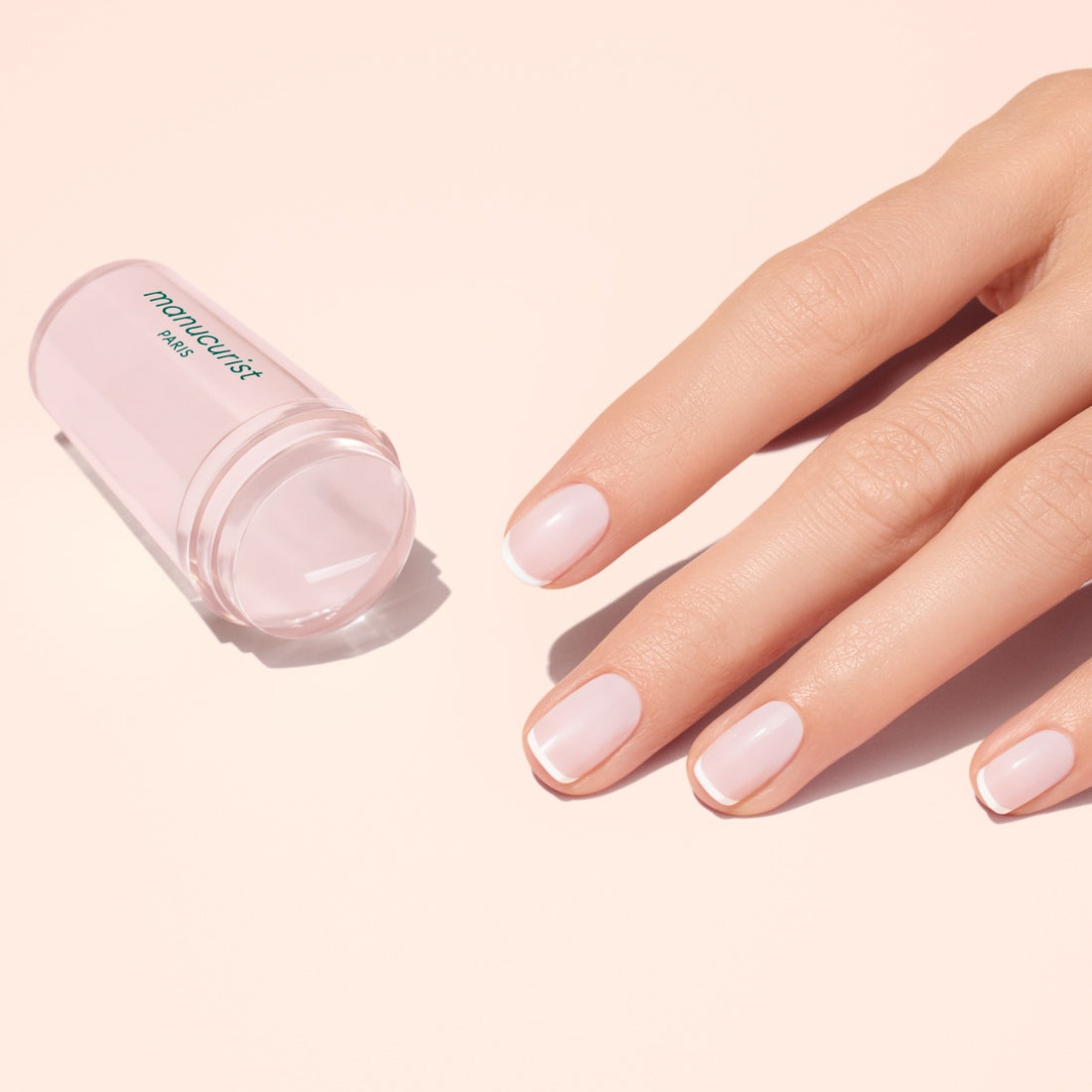 A hand with pale pink, glossy nails rests beside a translucent pink Mani stamper. The French Active™ Kit is perfect for creating flawless French tips at home.