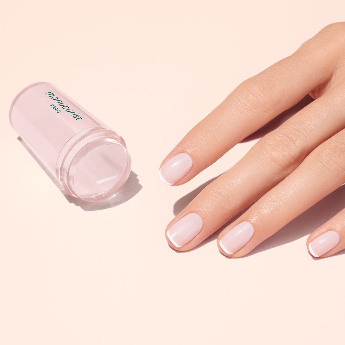 A hand with pale pink, glossy nails rests beside a translucent pink Mani stamper. The French Active™ Kit is perfect for creating flawless French tips at home.