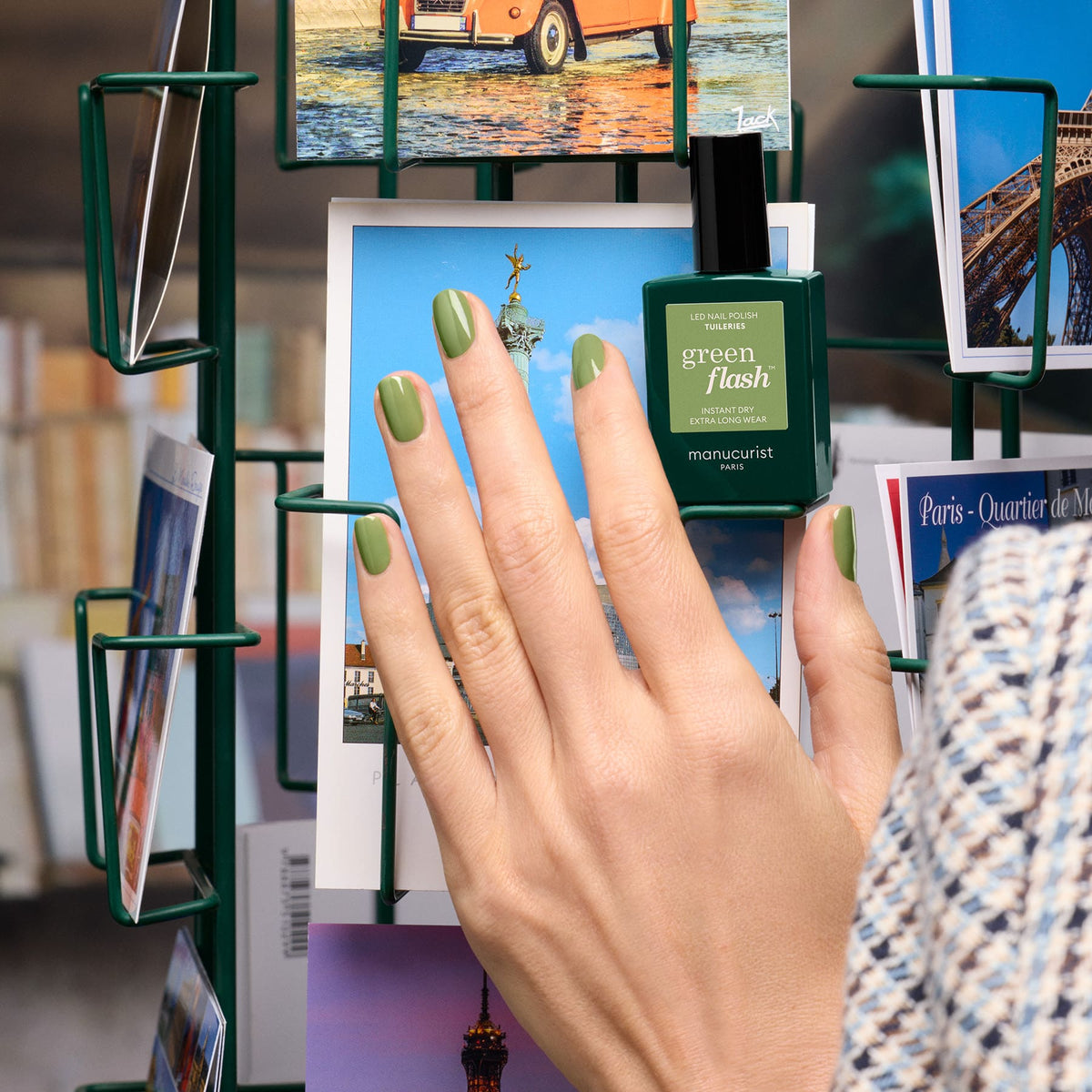 A hand with short, manicured nails painted olive green holds a Manucurist Tuileries Green Flash gel polish bottle in front of a rack of colorful postcards.
