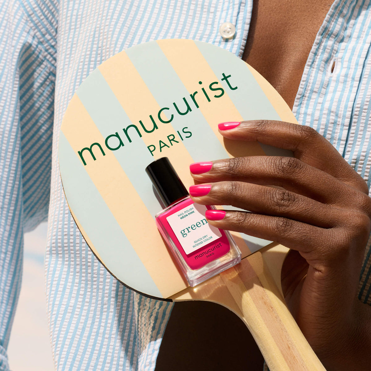 A person with Neon Pink nail polish by Manucurist holds a striped paddle labeled MANUCURIST PARIS and a matching bottle, wearing a blue and white striped shirt.