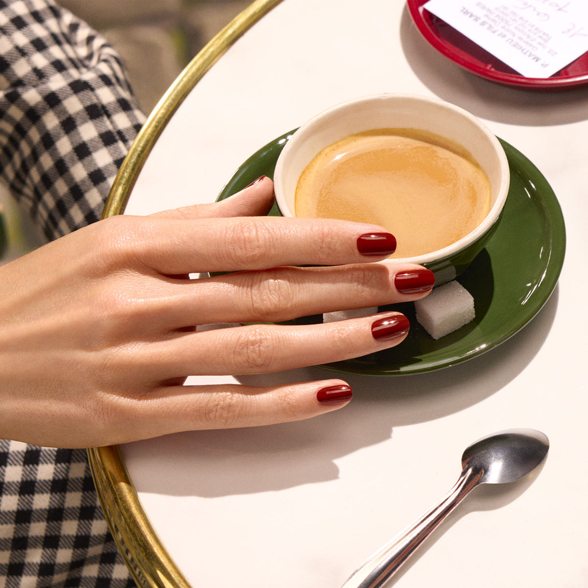 A hand with glossy Bohème polish by Manucurist rests on a table beside an espresso in a green saucer with two sugar cubes. A spoon and part of a check are seen, and the person wears black and white checkered clothing.