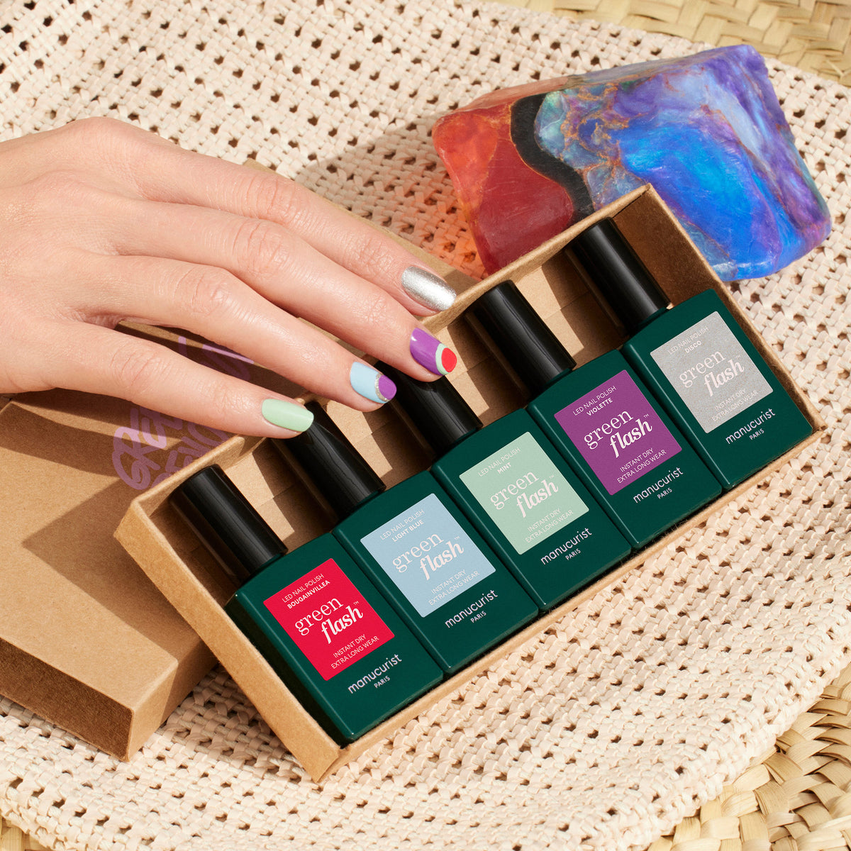 A hand with colorful Retro Rainbow nail art holds a Manucurist box of five Green Flash polishes. The box sits on a woven surface next to a vibrant translucent stone.
