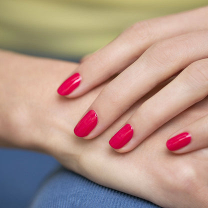 A close-up of a hand with neatly manicured nails painted in Peonie OLD by Manucurist-test, a bright pink 9-free nail polish, resting gently on another hand. The background is blurred with hints of beige and blue.