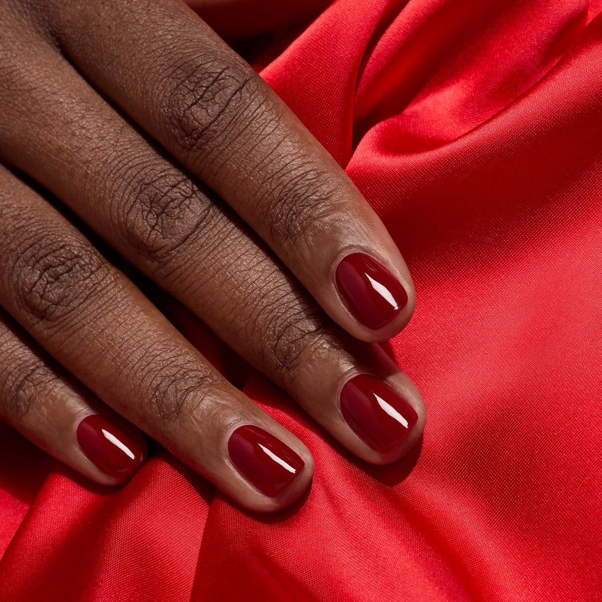 A close-up of a hand with neatly manicured nails painted in Manucurist’s Dark Pansy, resting on vibrant red satin fabric.
