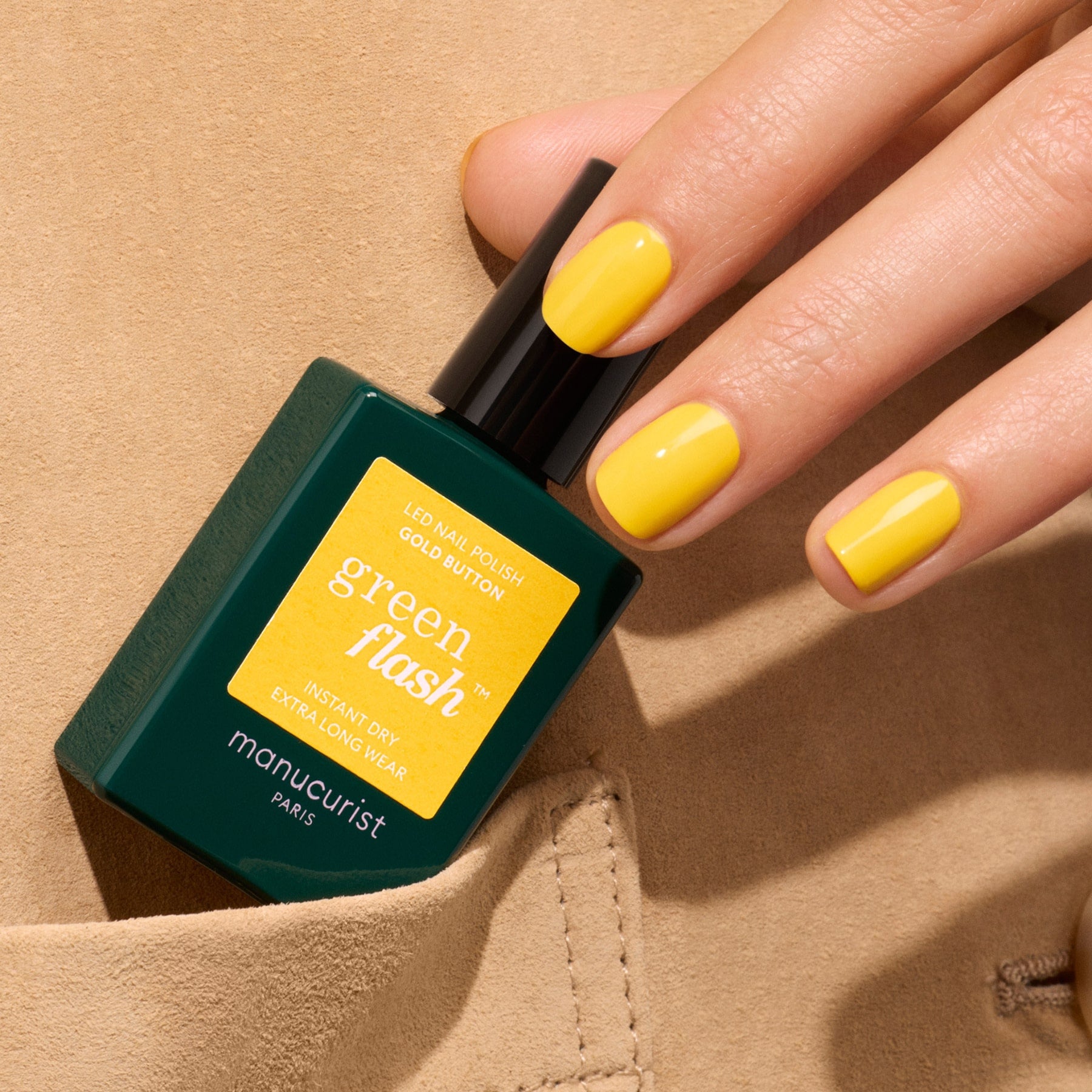 A hand with neatly manicured bright yellow nails holds a green bottle of Manucurist Gold Button polish, partially tucked into the pocket of a beige garment.