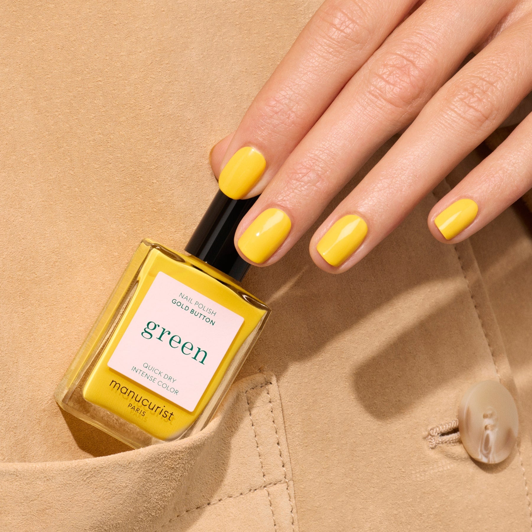 A hand with neatly manicured bright yellow nails holds a Gold Button nail polish bottle from Manucurist, resting on beige fabric with a button.
