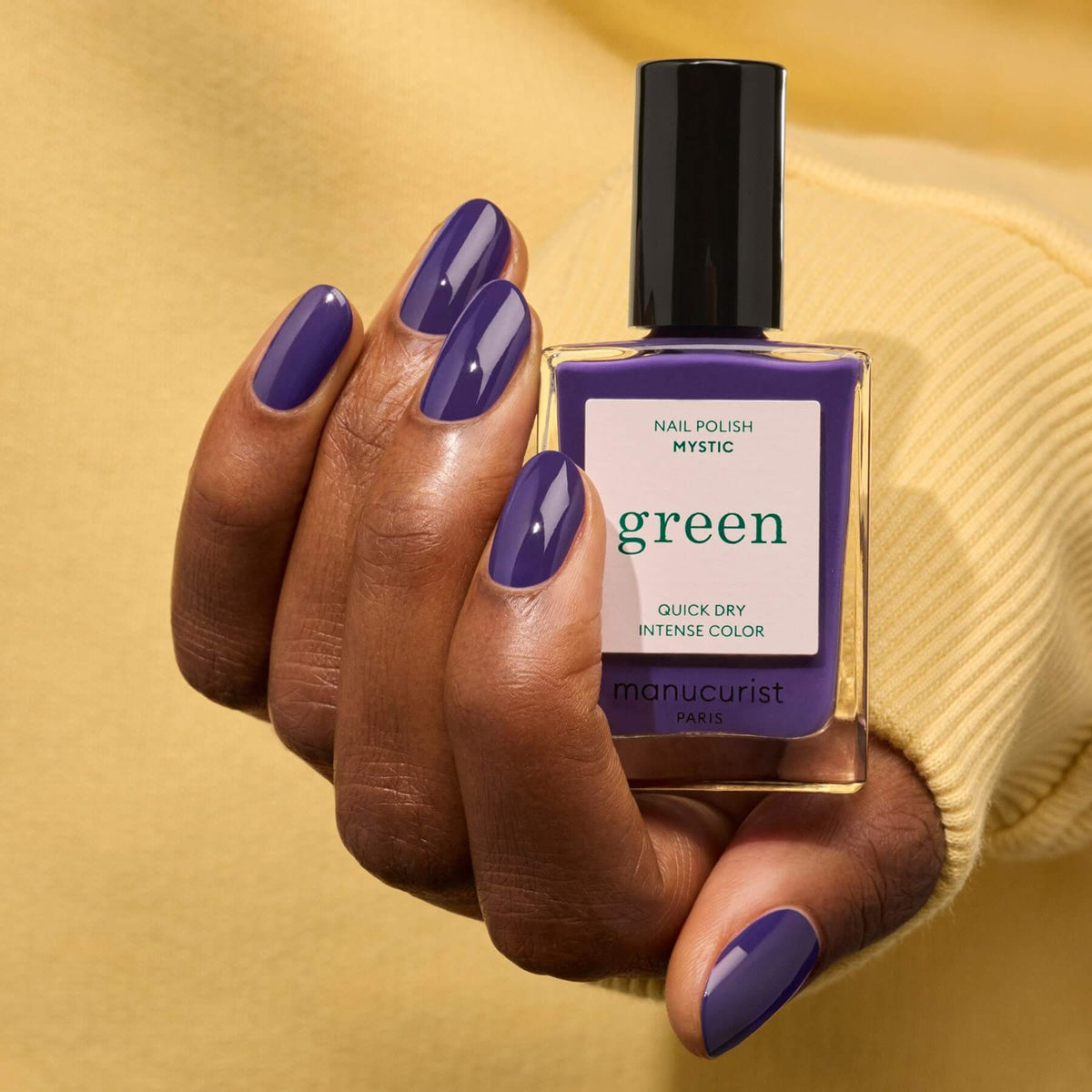 A hand with neatly manicured deep blue-violet nails holds a bottle of Manucurist Mystic nail polish against a yellow background.