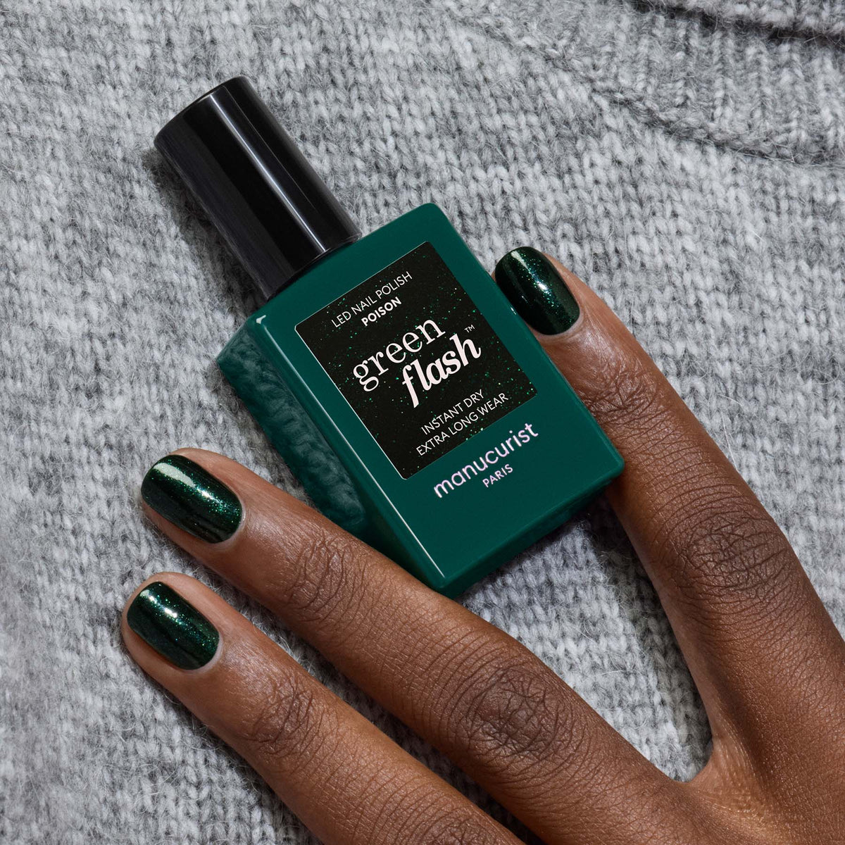 A hand with shiny forest green gel polish rests on gray knit fabric, holding a matching bottle of “Poison” LED nail polish by Manucurist.