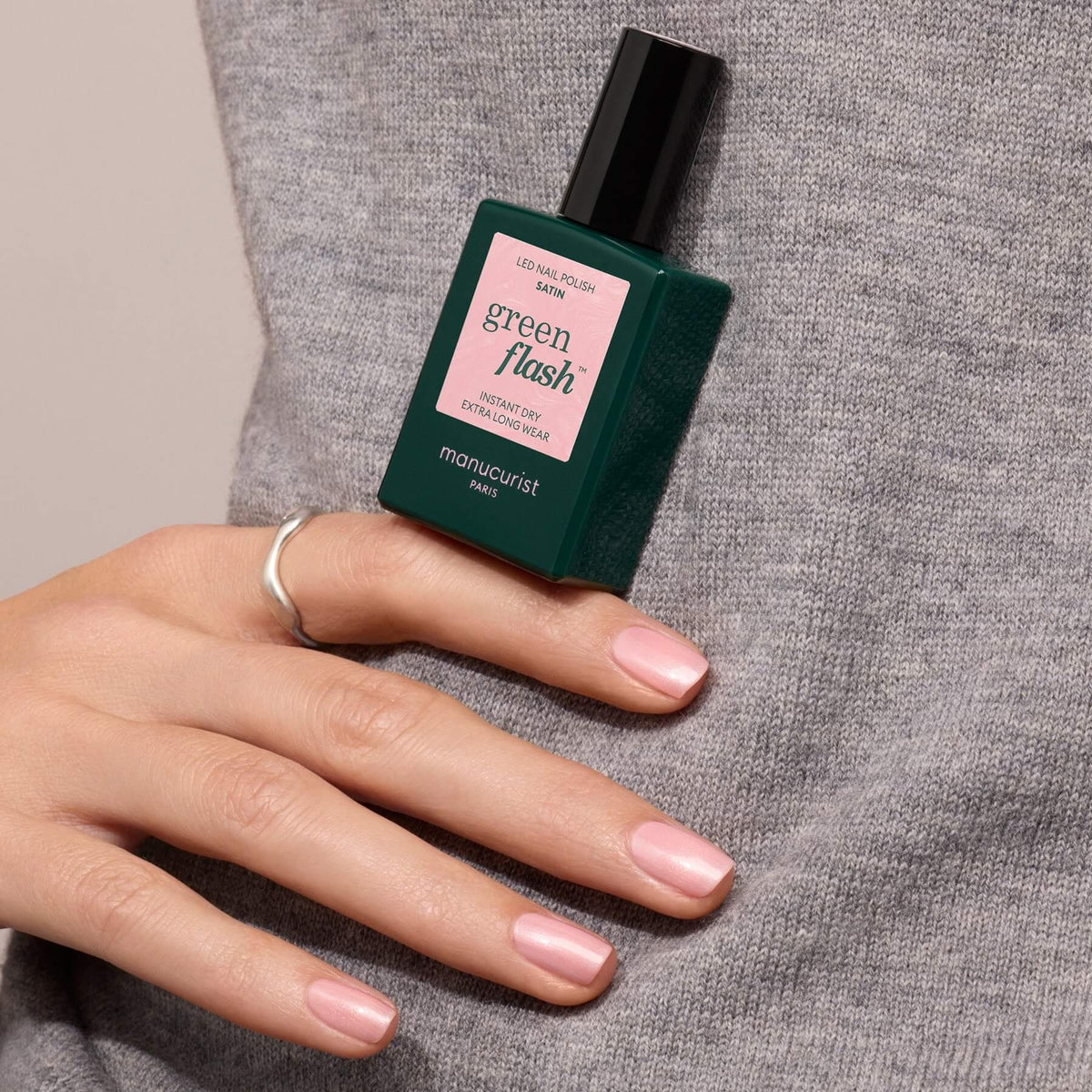 A hand with neatly manicured, pale pink nails rests on a gray sweater, holding a green bottle of Manucurist Satin polish for a semi-permanent finish.