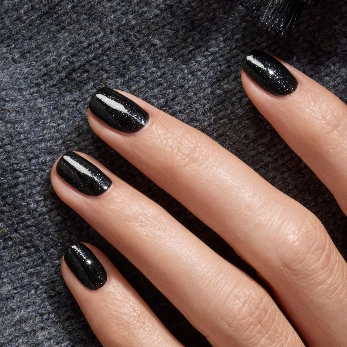 A hand with neatly manicured nails painted in Manucurist's Sparks, a shimmering black polish, rests on textured dark gray fabric.