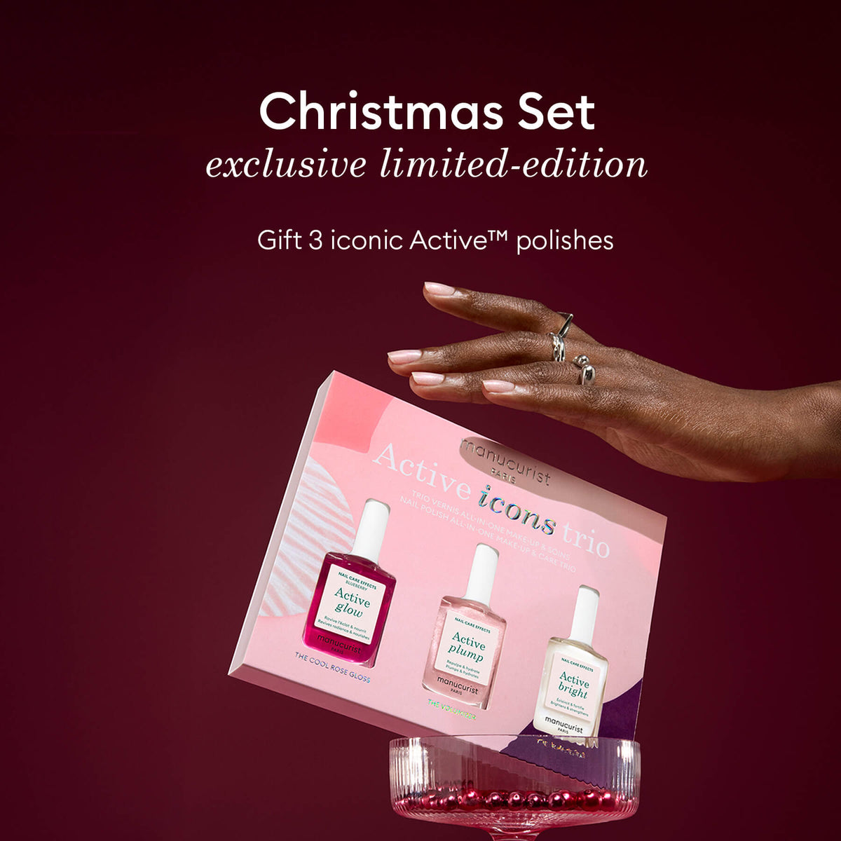 A hand holds a pink and white Manucurist Active Icons Trio box, featuring three exclusive limited-edition polishes—perfect as a Christmas nail polish gift set. A red dish with beads sits below.