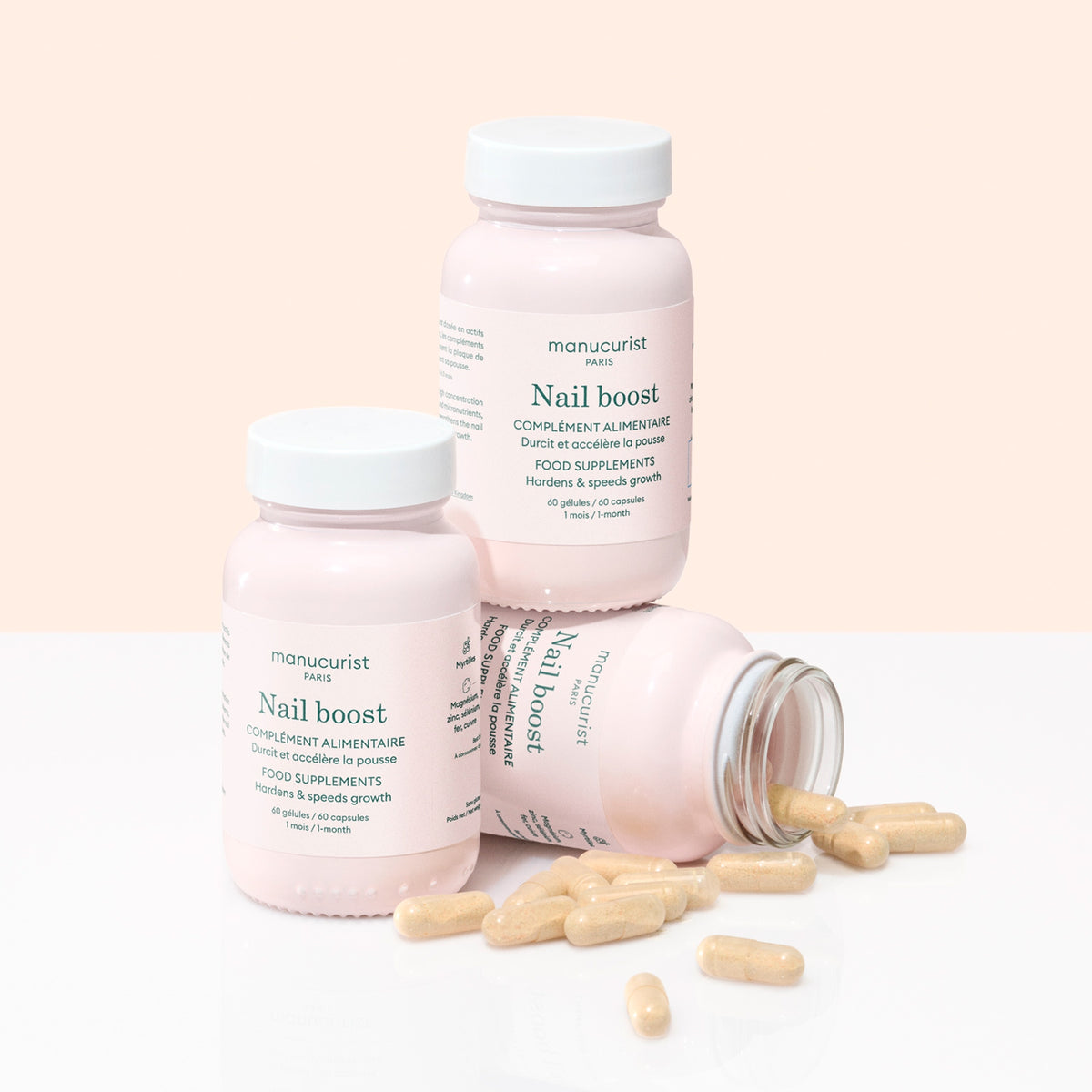 Three pink bottles of Manucurist Nail Boost are arranged against a pale pink background, with one lying on its side spilling yellow nail supplements and two standing upright, designed to help strengthen nails.