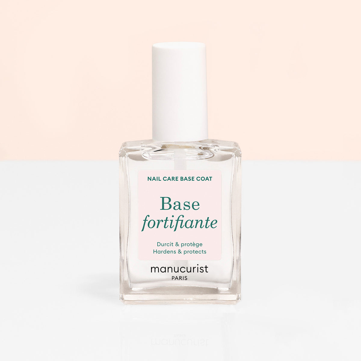 A clear glass bottle of Manucurist Force Nail Hardener with a white cap, set against a pale peach and white background. The label notes this nail treatment is formulated to harden and strengthen nails.
