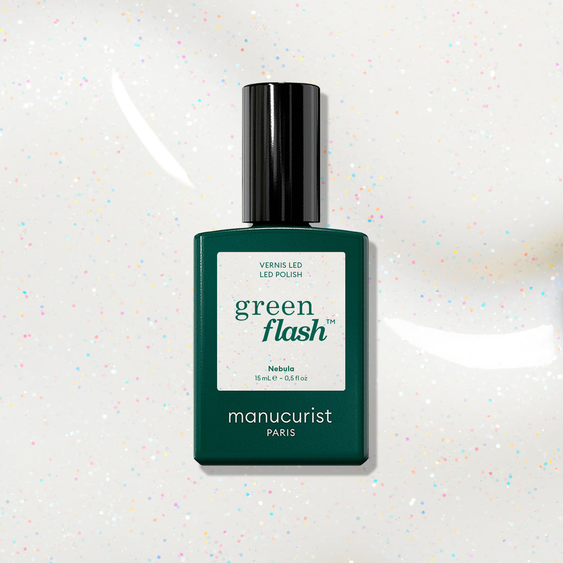 A dark green Manucurist bottle of Green Flash LED polish in shade Nebula is displayed on a white background, showing holographic glitter specks.