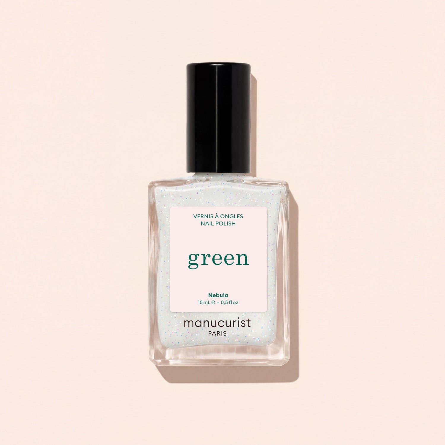 A clear glass bottle of Manucurist nail polish in the shade Nebula, featuring a black cap and white label, is displayed on a pale beige background.
