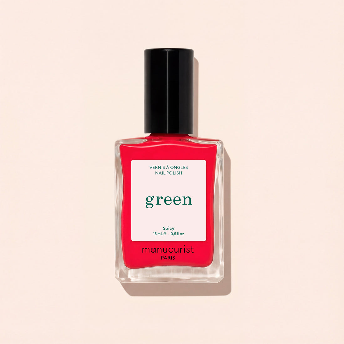 A clear glass bottle of Manucurist nail polish in the Spicy shade, featuring a black cap and “green” labeling, rests on a light beige background.