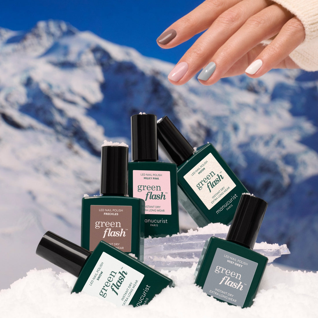A hand with multicolored manicured nails reaches above several bottles of Manucurist’s Rainbow Collections, arranged in snow against a snowy mountain backdrop, showcasing curated winter colour palettes for stylish LED gel manicures.
