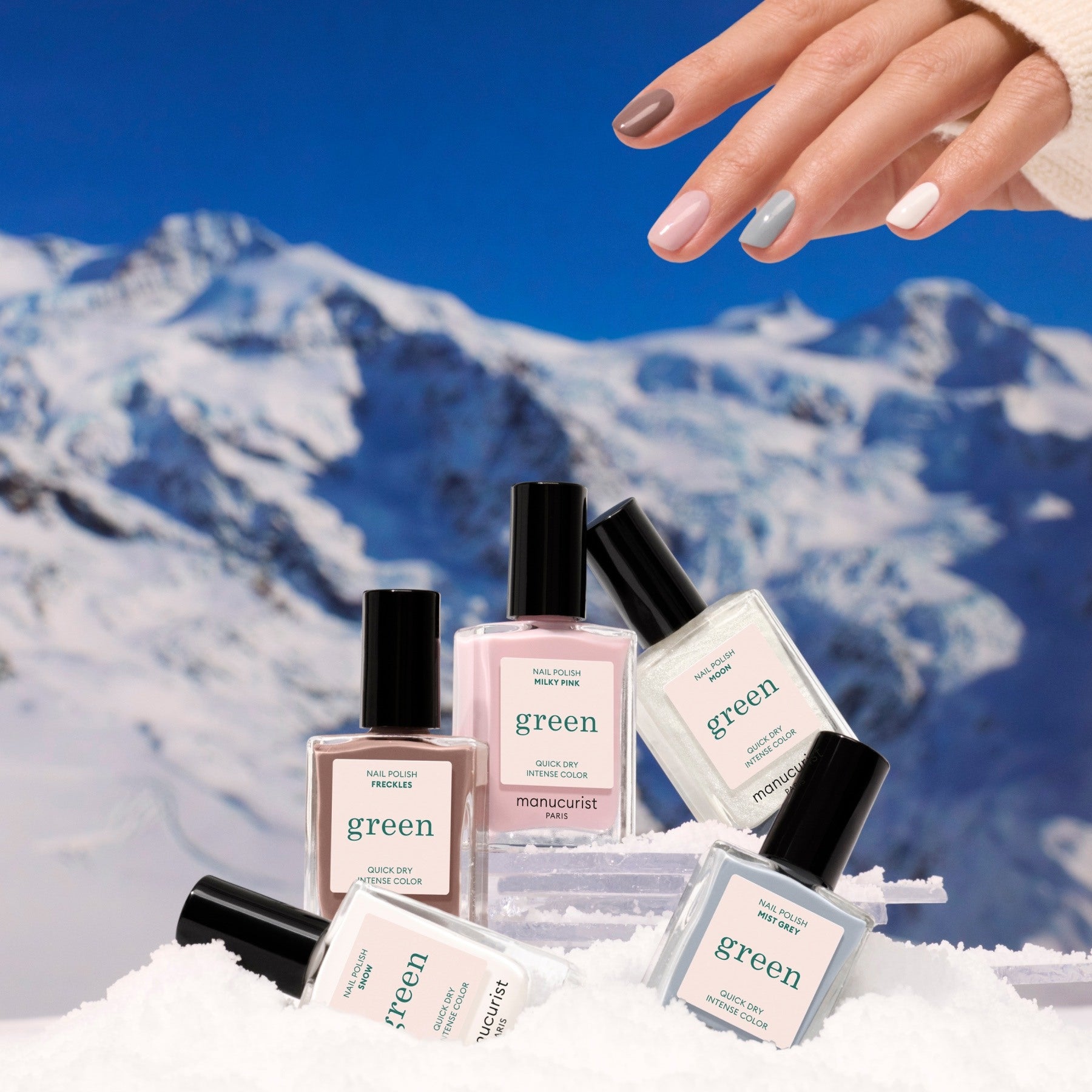 A hand with multicolored nails hovers above five bottles of Manucurist Rainbow Collections nail polish on snow, set against a snowy mountain scene—highlighting curated shades inspired by the latest manicure trends.