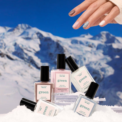 A hand with multicolored nails hovers above five bottles of Manucurist Rainbow Collections nail polish on snow, set against a snowy mountain scene—highlighting curated shades inspired by the latest manicure trends.