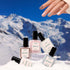 A hand with multicolored nails hovers above five bottles of Manucurist Rainbow Collections nail polish on snow, set against a snowy mountain scene—highlighting curated shades inspired by the latest manicure trends.