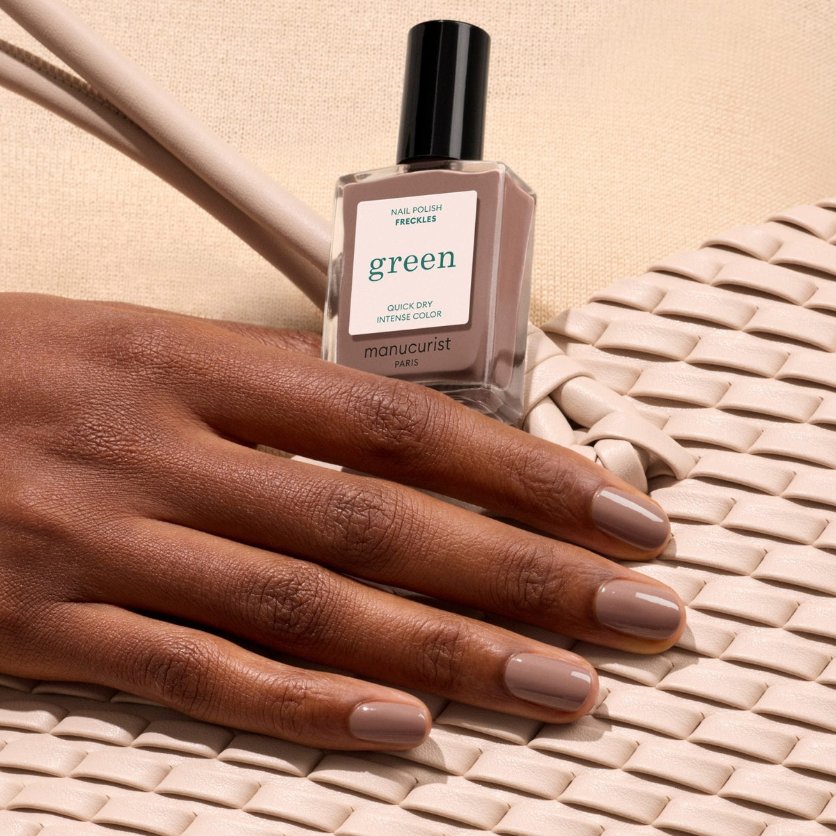 A hand with neatly manicured, wood-toned brown nails holds a bottle of Manucurist “Freckles” nail polish, resting on a white woven bag against a beige textured fabric background.