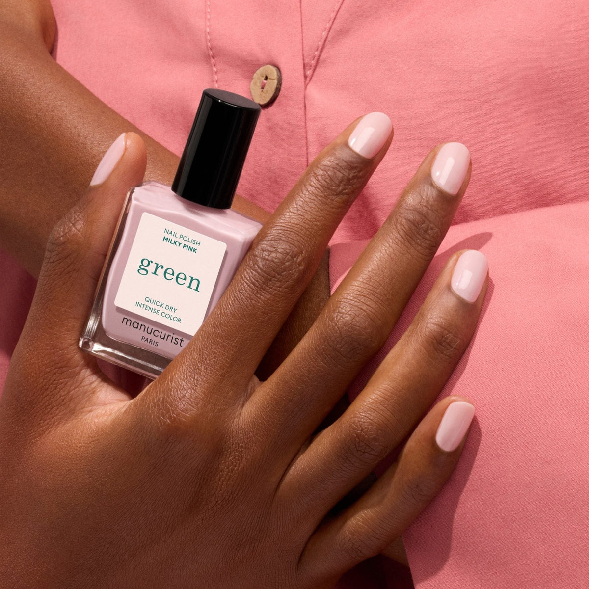 A hand with neatly manicured Milky Pink nails holds a Manucurist bottle. The person wears a pink button-up shirt, highlighting the Milky Pink polish, Manucurist branding, and their stylish outfit.