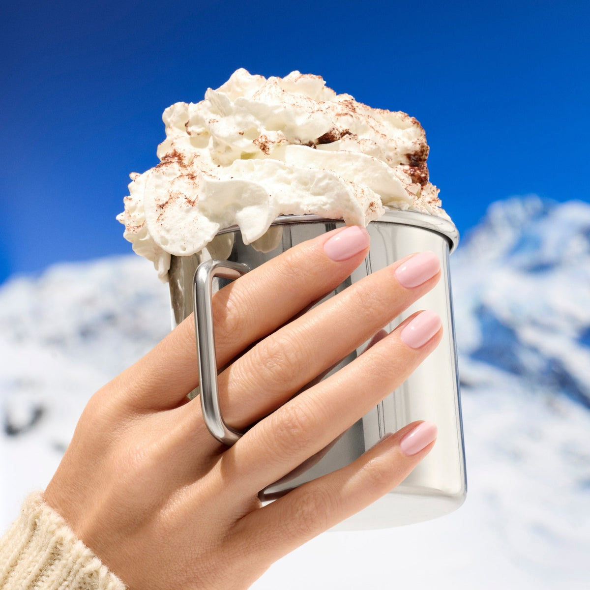 A hand with Manucurist’s Milky Pink gel polish holds a metal mug topped with whipped cream and cocoa, set before snowy mountains beneath a clear blue sky.