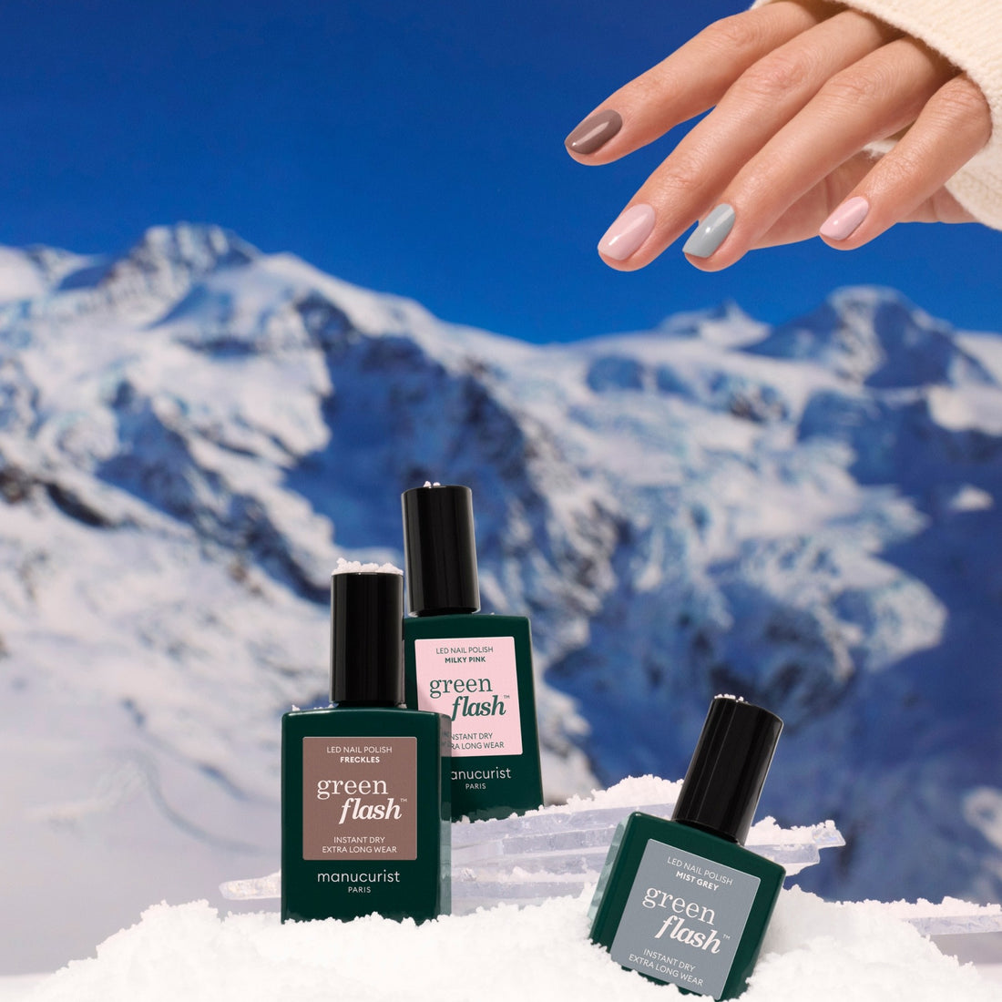 A manicured hand with nude and gray nails, inspired by the latest trends, holds three green bottles of Manucurist Trio Collections nail polish on snow with snowy mountains in the background.