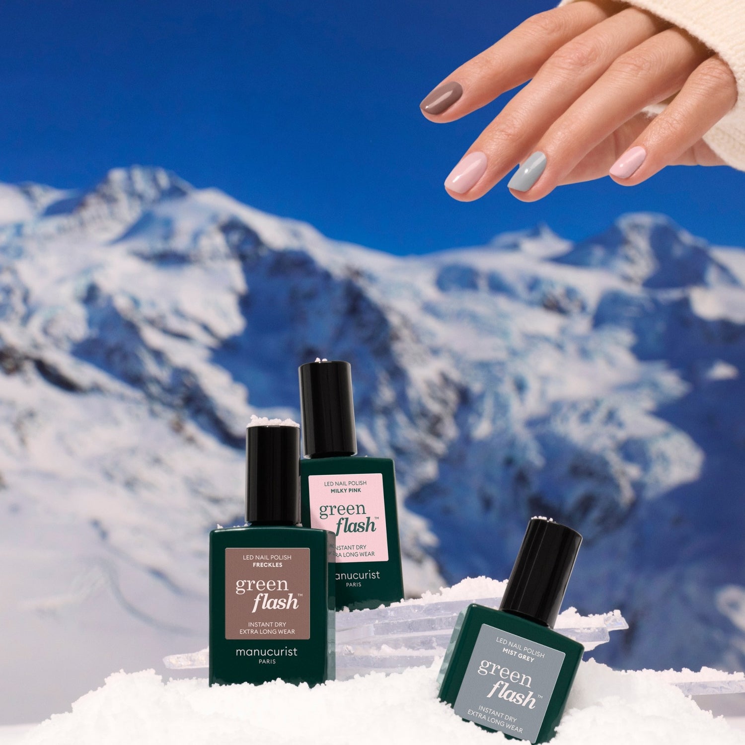 A manicured hand with nude and gray nails, inspired by the latest trends, holds three green bottles of Manucurist Trio Collections nail polish on snow with snowy mountains in the background.