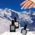 A manicured hand with nude and gray nails, inspired by the latest trends, holds three green bottles of Manucurist Trio Collections nail polish on snow with snowy mountains in the background.