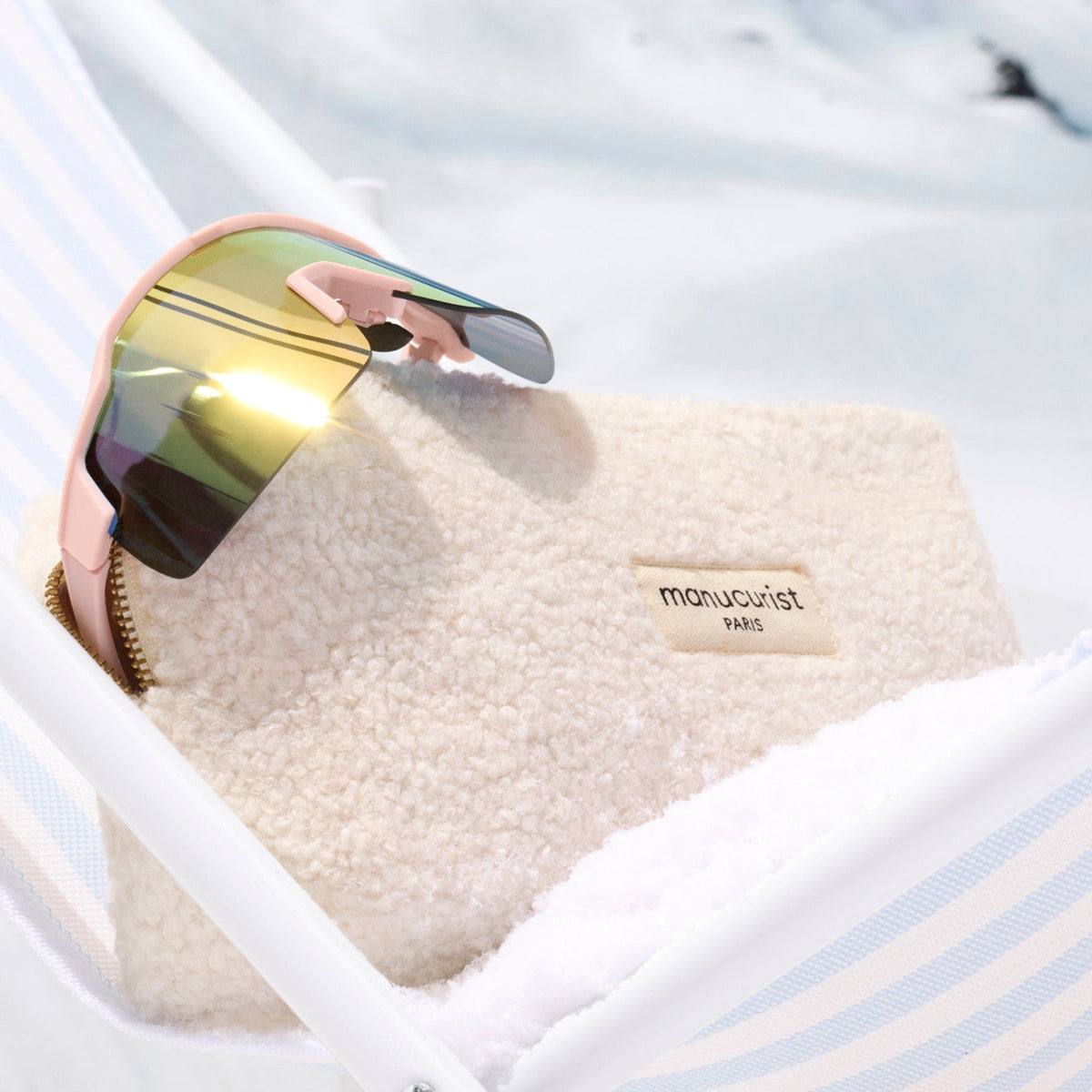 A pair of sunglasses with reflective lenses rests on the Teddy Pouch by Manucurist, set atop striped fabric with a snowy background.