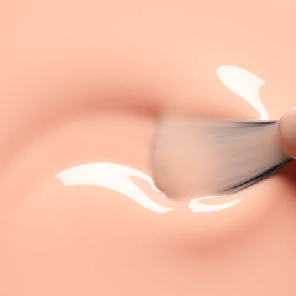 A close-up of a makeup brush applying a glossy, creamy substance onto smooth, peach-colored skin evokes the soft radiance of Manucurist’s Milky Peach gel polish.