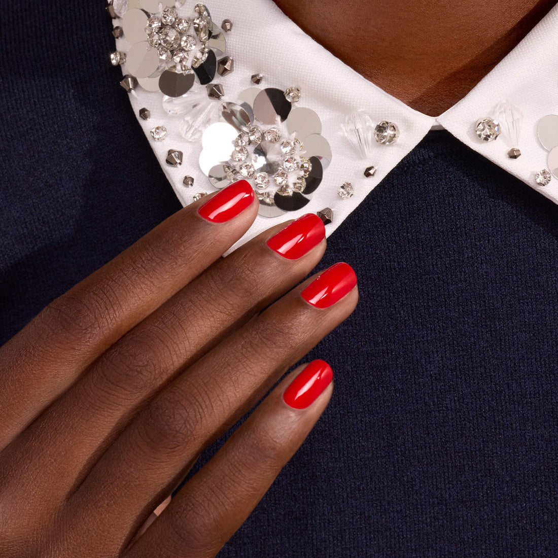 A hand polished with Manucurist’s Anémone, a glossy red shade with subtle blue undertones, rests on dark fabric. The person wears a white collar embellished with rhinestones and sequins, adding a sparkling, elegant touch to the look.