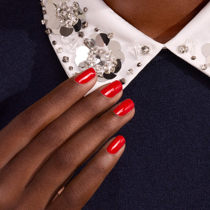 A hand polished with Manucurist’s Anémone, a glossy red shade with subtle blue undertones, rests on dark fabric. The person wears a white collar embellished with rhinestones and sequins, adding a sparkling, elegant touch to the look.