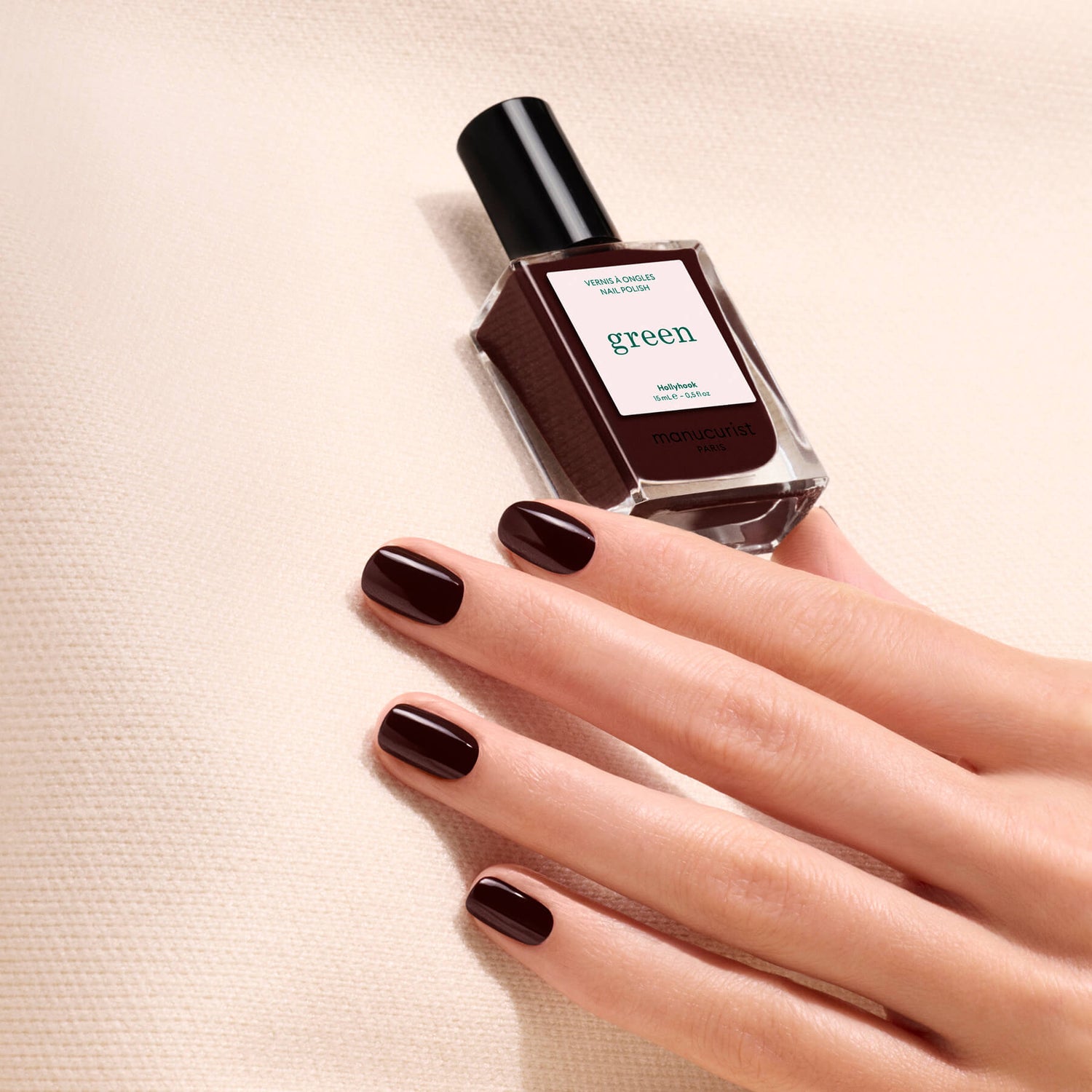 A hand with neatly manicured, dark brown nails holds a bottle of Manucurist Hollyhock nail polish against a beige textured background.