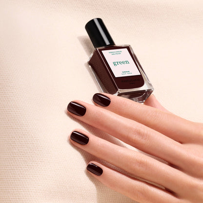 A hand with neatly manicured, dark brown nails holds a bottle of Manucurist Hollyhock nail polish against a beige textured background.