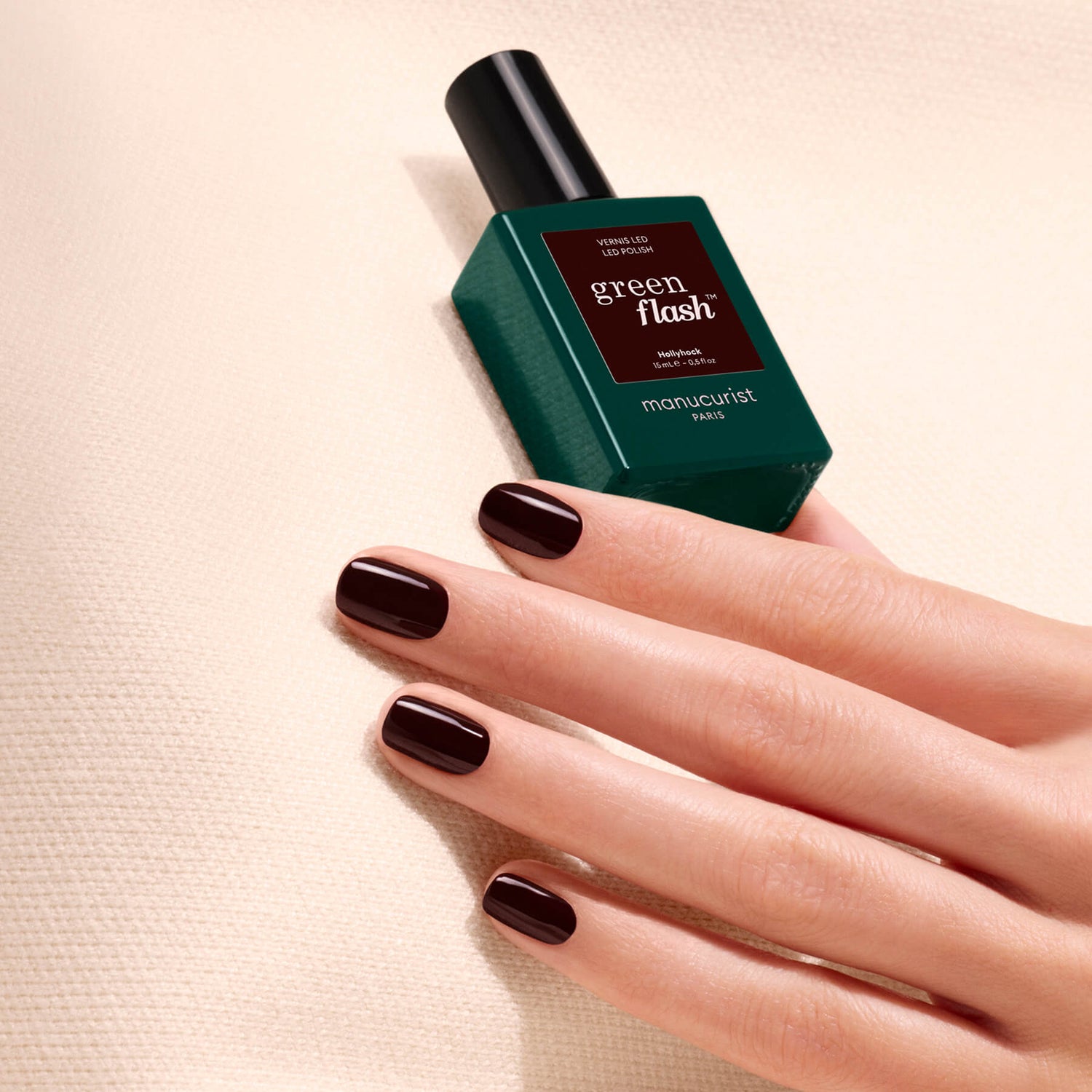 A hand adorned with dark, glossy nail polish—reminiscent of a deep red shade called Hollyhock by Manucurist—rests on a textured cream surface next to a green bottle of Green Flash gel polish.