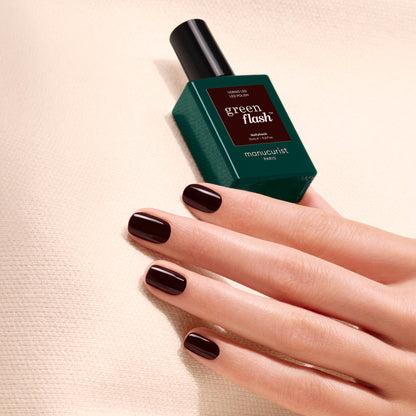 A hand adorned with dark, glossy nail polish—reminiscent of a deep red shade called Hollyhock by Manucurist—rests on a textured cream surface next to a green bottle of Green Flash gel polish.