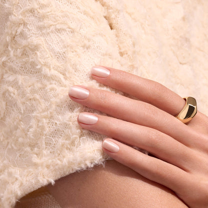 A hand with light pink polished nails, enhanced by Manucurist’s Pearlescent nail polish, rests on textured cream-colored fabric. The person is wearing a large, modern gold ring on their finger.