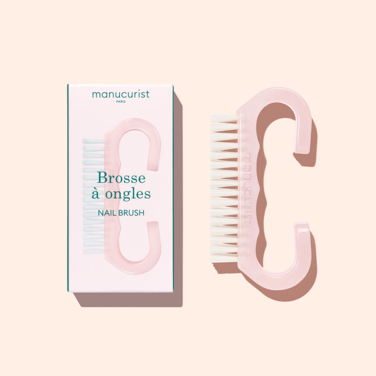 A pink Nail Brush from Manucurist, featuring vegan bristles, is displayed beside its box labeled “Manucurist Paris, Brosse à ongles, Nail Brush.” Ideal for an at-home manicure, both the brush and its box are set against a pale peach background.