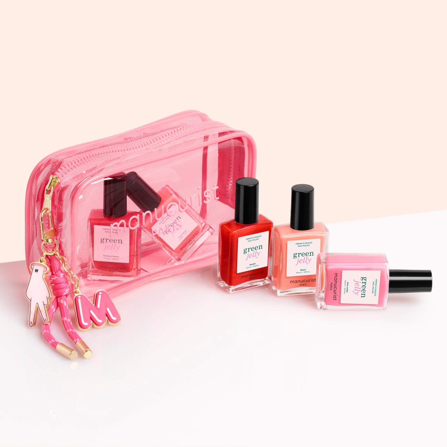 Three Manucurist Jelly Charm nail polishes in red, orange, and pink sit before a transparent pink zippered pouch, accented with a keychain featuring a pink "M" and Jelly Charm details.