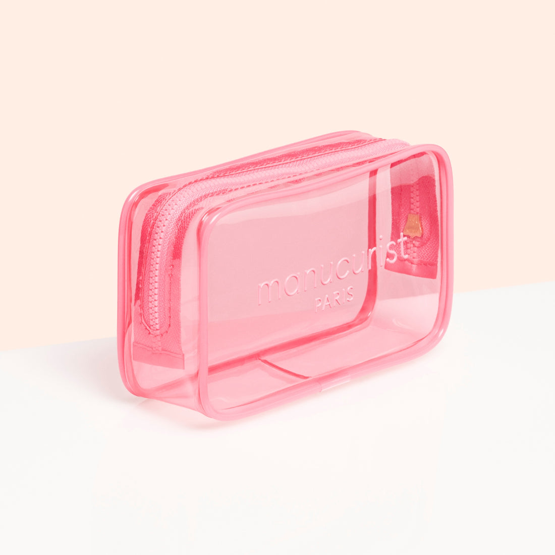 The Manucurist Mini Jelly Bag is a small, rectangular, transparent pink bag with a zipper and the brand logo embossed on the front, ideal as a travel manicure bag. Displayed against a clean white and light peach background.