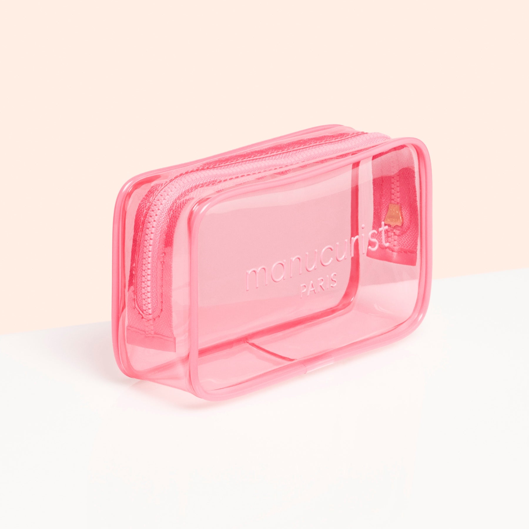 The Manucurist Mini Jelly Bag is a small, rectangular, transparent pink bag with a zipper and the brand logo embossed on the front, ideal as a travel manicure bag. Displayed against a clean white and light peach background.