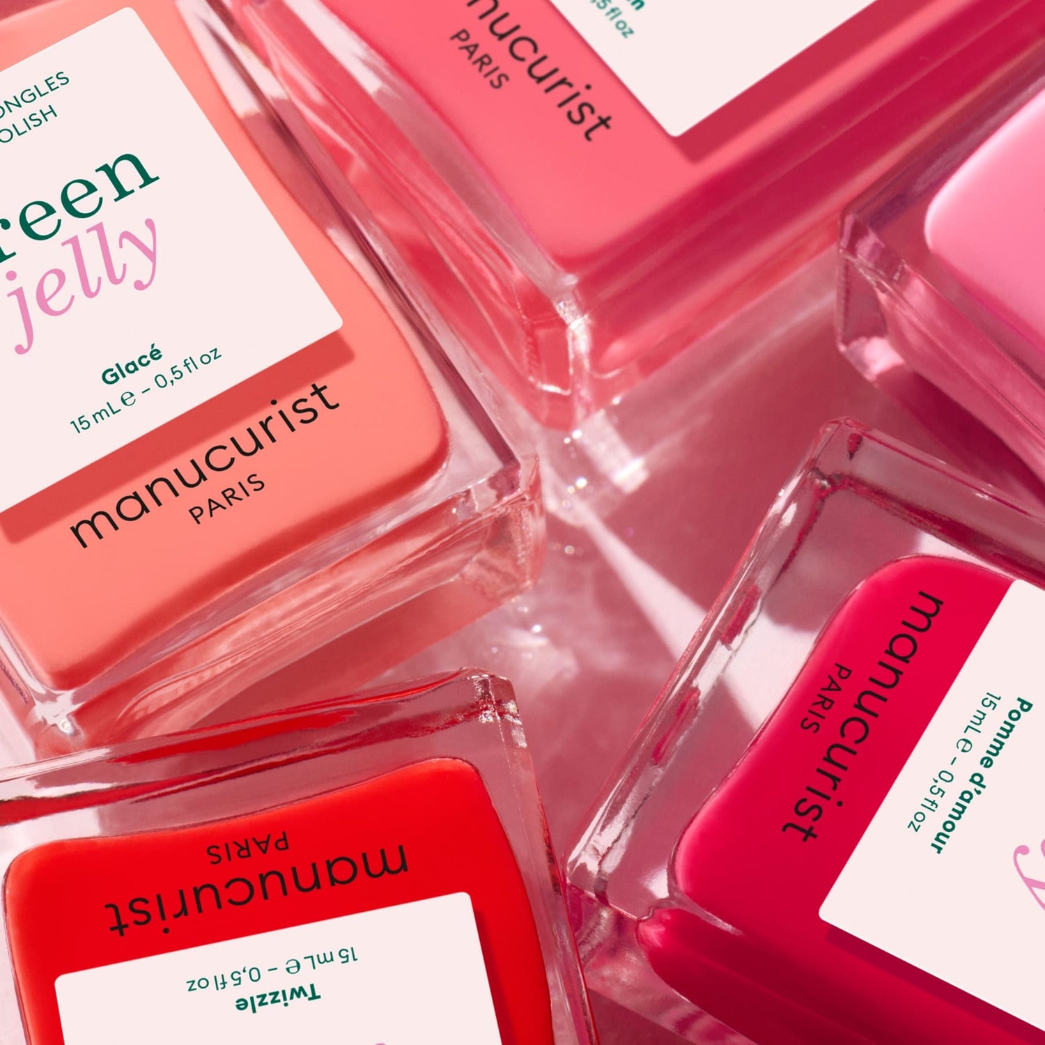 Close-up of several bottles of Manucurist Glacé ultra-glossy nail polish in pink, coral, and red shades, arranged diagonally on a light pink surface, with labels showing product names and details visible.