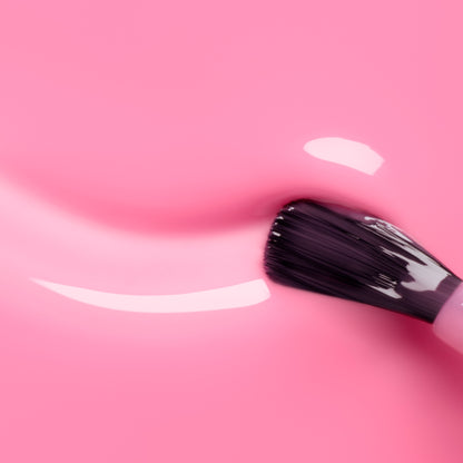 A close-up of a brush applying Manucurist Pink Sorbet Jelly, a translucent pink nail polish, on a smooth pink surface, with reflections showcasing its shiny texture.
