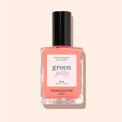 A bottle of Manucurist Glacé ultra-glossy jelly nail polish in coral pink with a black cap, shown against a light peach background.