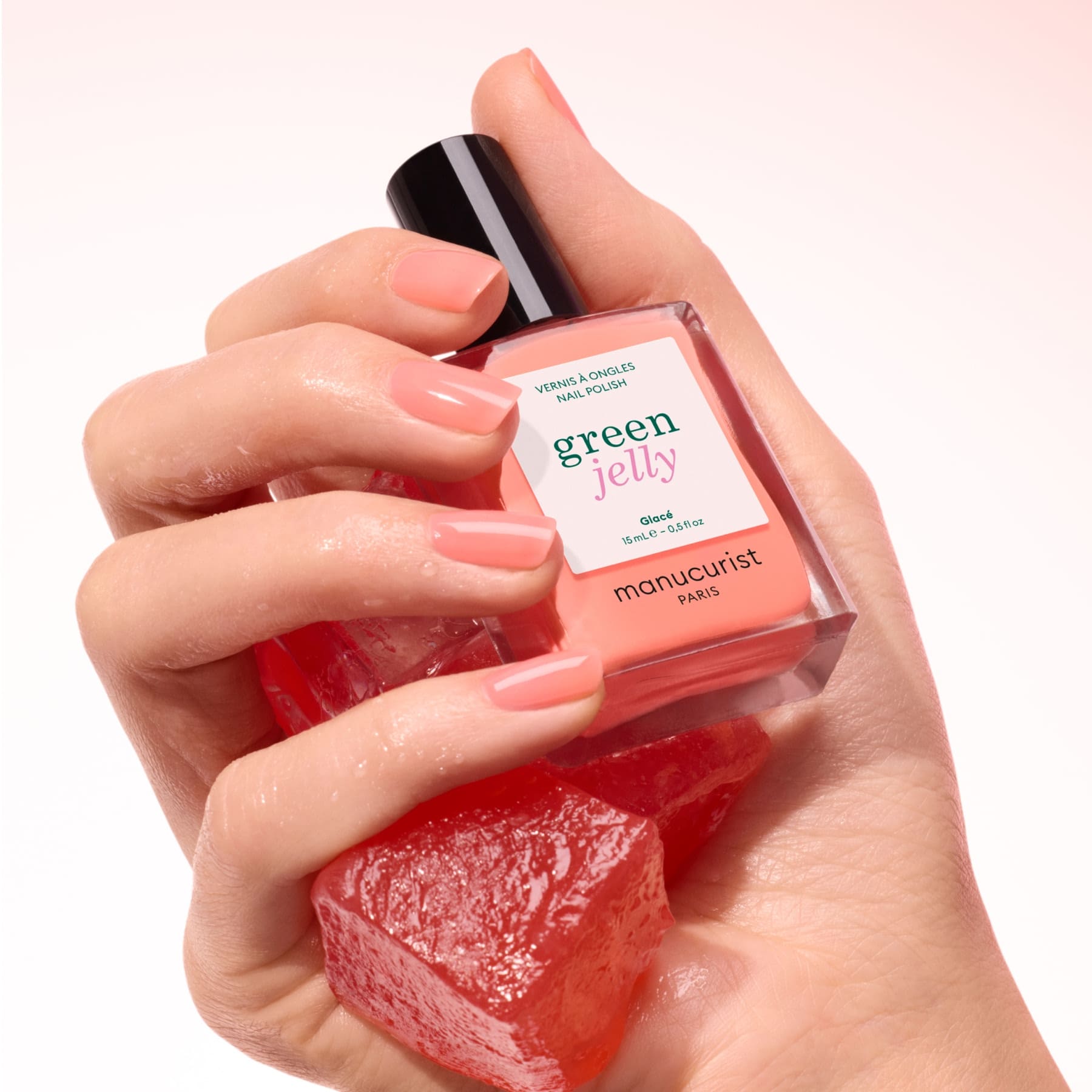 A hand with ultra-glossy, peach-colored nails holds a bottle of Manucurist Glacé and a red translucent gel, set against a soft pink background.