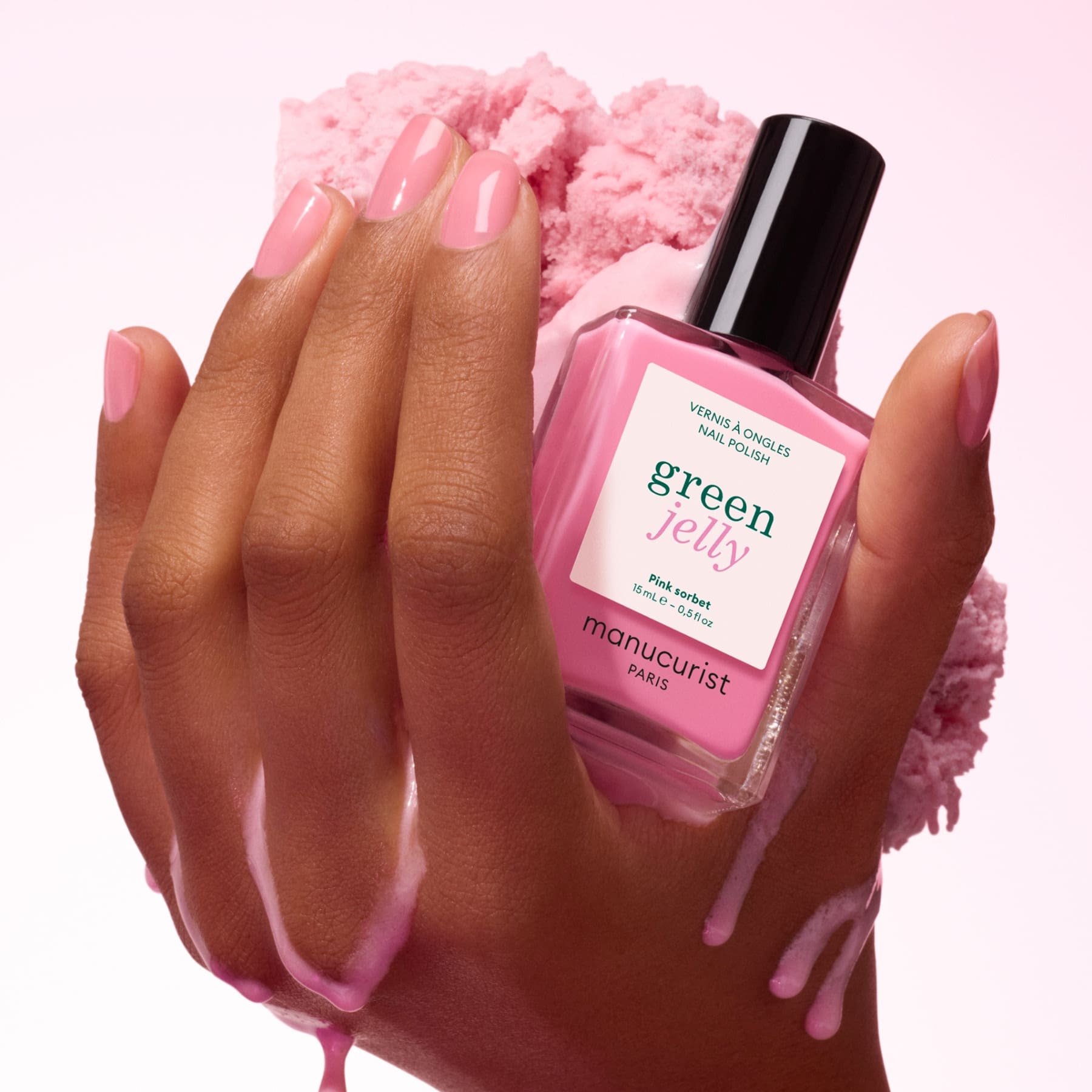 A hand with ultra-glossy pink nails holds a bottle of Pink Sorbet Jelly nail polish by Manucurist, with pink liquid dripping down the fingers against a textured pink background.