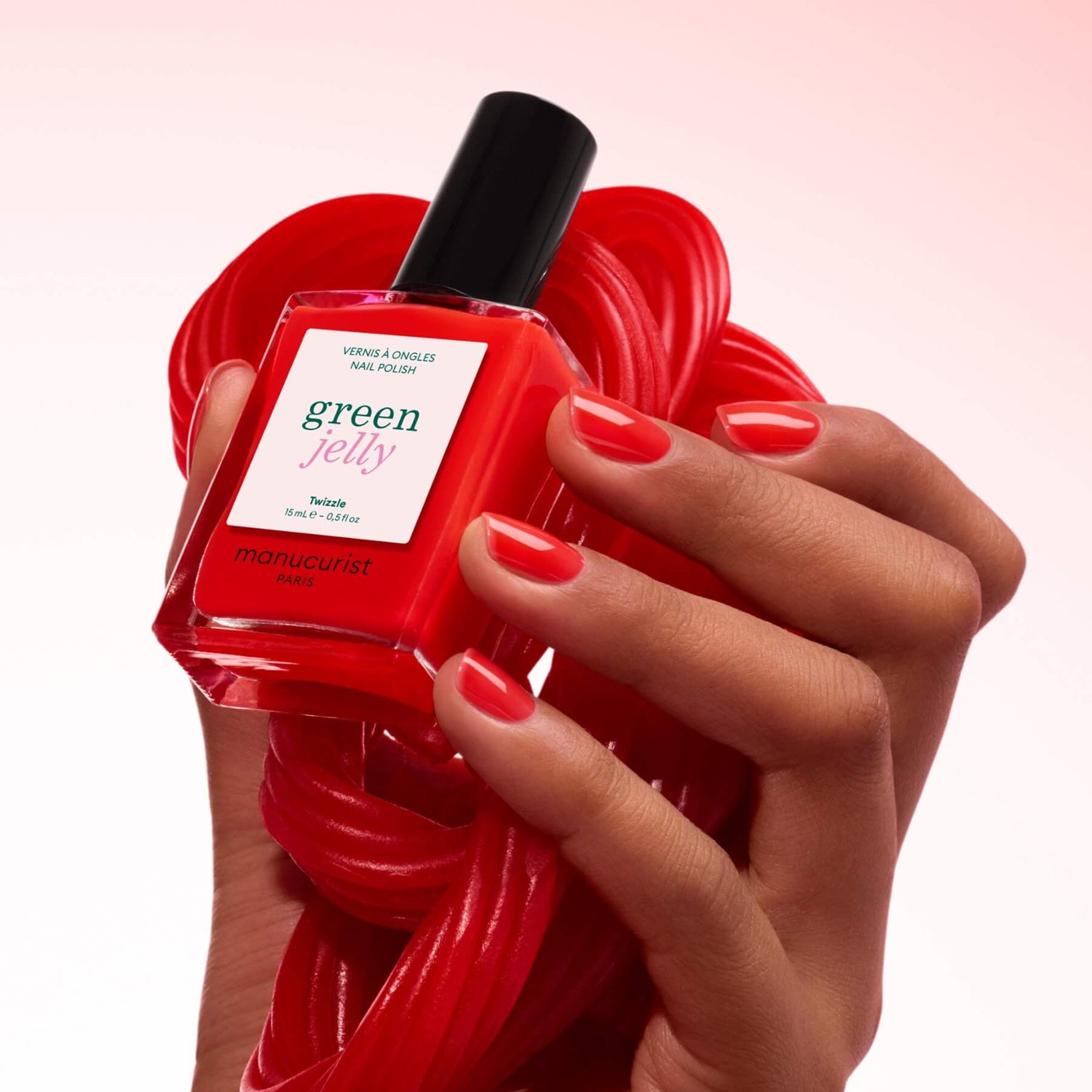 A hand with neatly painted red nails holds a bottle of Manucurist Twizzle polish in a vibrant red shade, with a coiled red candy in the background.