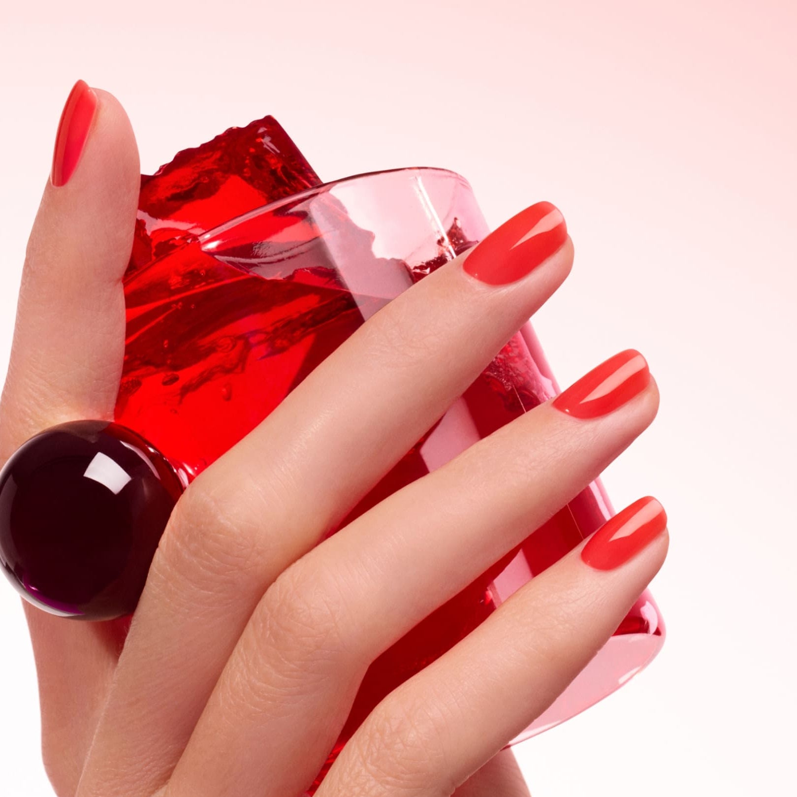 A hand with ultra-glossy, red-orange Twizzle nails by Manucurist holds a translucent red object and a clear glass, wearing a large, round dark ring against a soft gradient of pale pink to white.